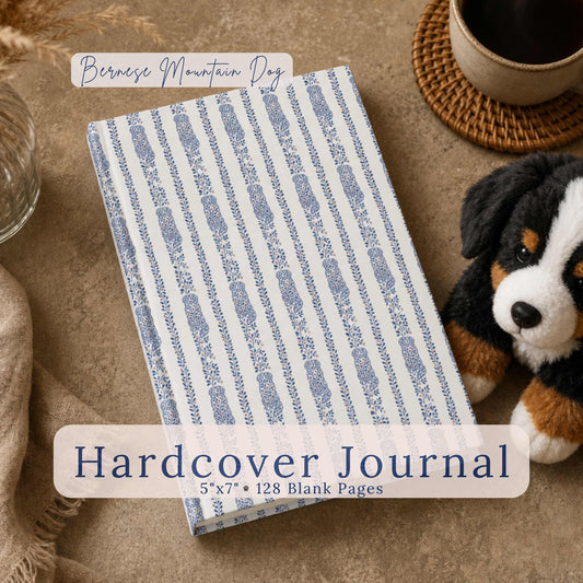 A Bernese Mountain Dog plush toy sits beside a hardcover journal featuring a delicate blue vine-and-leaf stripe with a Bernese Mountain Dog silhouette woven throughout on white, with a circular close-up inset of the pattern, on a warm taupe surface with dried pampas grass and a coffee cup. Text reads "Bernese Mountain Dog Hardcover Journal, 5"x7" • 128 Blank Pages."